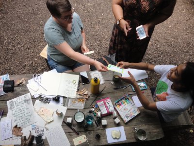 image from above of a craft table on woodchip ground, Linden McMaoh and Shair Begum pass an artwork between them whilst someone else nearby stands with a smartphone and pointed finger 