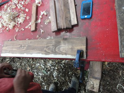 a piece of wood clamped to a table with wood shavings surrounding it. A smartphone is also visible along with feet and hands in motion