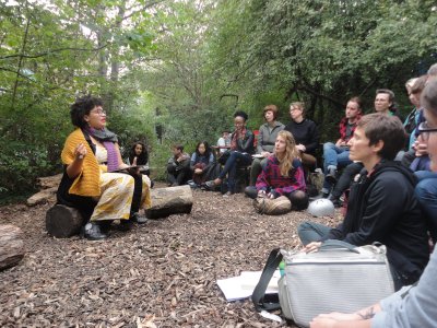 Ama Josephine Budge sits on a lot in a nature reserve, gesticulating with one hand whilst reading to an audience sitting on the ground and on seats