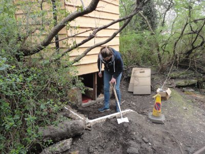 a person digs with a spade, white pipe appears under the ground surrounded by gravel, open empty compost toilet chambers in the background with a 'caution' building cone to the side