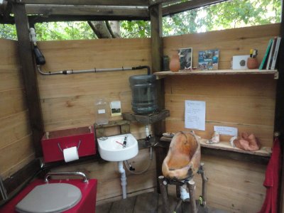 interior of compost toilet shows pink toilet box, basin, rainwater collection tank, shelves with books and pots on and an orangey vessel which is an all-gender urinal / 'liquid loo'