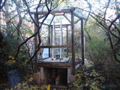 raised open wooden structure under construction in wooded setting with autumn leaves on the ground, a clear polycarbonate roof is being attached, and a ladder is standing on the floor. Under the structure are two brick chambers with a low brick wall separating them, and a blackboard which reads "compost toilet jobs" followed by a list