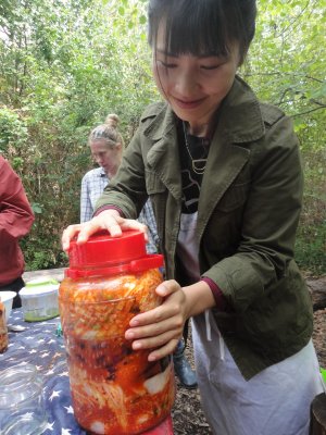 large pot of red / orange kimchi held lovingly by Nayoung Jeong, 2 people are standing in the background