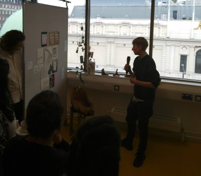 Hari speaks to mobile handing over all-gender urinal, holding a microphone. They are standing against a window, with an audience listening