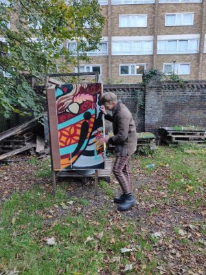 Colourful toilet cubicle with graffiti designs on the walls and door stands in an outdoor setting with trees and flats nearby, hari is in movement, adjusting the door