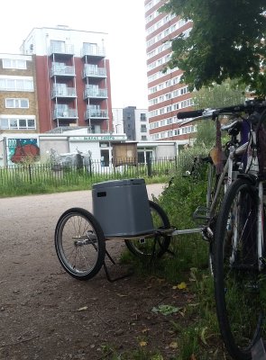 grey plastic camping toilet on the back of a bike trailer with Soanes Centre and a block of flats in the background