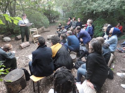 Linden Mcmahon reads from a notebook with around 20 audience members sitting around, with trees and logs in the surroundings 