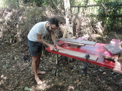 someone with bare feet and tattoos planes a plank of wood on a red table in a wooded setting