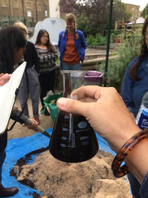 hand holds up chemistry beaker containing dark brown soil solution, in the background a group is gathered around a pile of bran and leaf mould on a blue tarp, which someone mixes with a spade