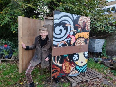 Hari wearing brown coat and chequered trousers appears coming out of the toilet with one leg extended and a large grin on their face