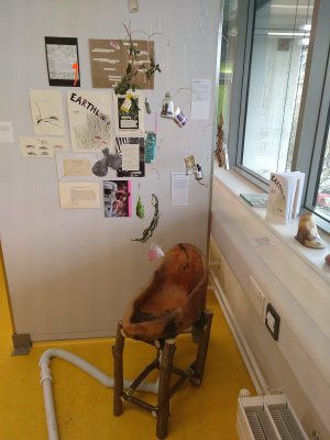 mobile dangles over an earthenware all gender-urinal with pipes attached. In the background is a collage of zine pages, with a zine presented on a nearby windowsill